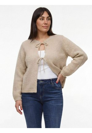 Nő bézs kötött kardigánban, amelyet két masnival kötöttek át, fehér felső és kék farmer nadrág fölött, kezét csípőjén tartva, fehér háttér előtt pózol.