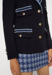 Blazer noir texturé avec bordure bleu clair et boutons dorés, assorti à une mini-jupe à motif pied-de-poule bleu et blanc.