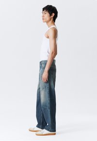 Nuori mies, joka on pukeutunut löysiin kaksivärisiin sinisiin farkkuihin, valkoiseen hihattomaan paitaan ja valkoisiin lenkkareihin, seisoo profiilissa yksinkertaista valkoista taustaa vasten.