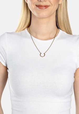 Guldfarvet halskæde med en cirkulær vedhæng på en tynd kæde, båret over en tætsiddende hvid t-shirt med korte ærmer. Enkel og moderne design.