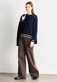 Μπλε πουλόβερ με λευκές ρίγες στο τελείωμα και τις μανσέτες, χρυσά κουμπιά στον έναν ώμο, σε συνδυασμό με καφέ παντελόνι φαρδύ και μαύρα παπούτσια.