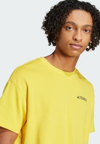 Gul bomull T-skjorte med avslappet passform og korte ermer, med en svart TERREX-logo på brystet. Jevn tekstur og rund hals.