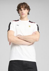 Camiseta de algodón blanca con mangas negras y acentos rojos, que presenta un logo de Puma y un emblema de Ferrari en el pecho. Diseño de ajuste regular.