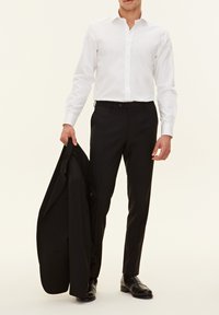 Camicia bianca con bottoni e maniche lunghe, abbinata a pantaloni neri. Tiene in mano un giacca da completo nera. Scarpe da vestir nere.