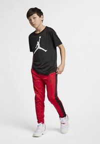 Gutt som står med svart Jordan t-skjorte, rød og svart treningsbukser, og hvite sneakers foran en ren hvit bakgrunn.