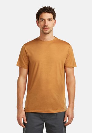 Mężczyzna o krótkich kręconych włosach, ubrany w prostą brązową koszulkę z krótkim rękawem i ciemne spodnie, stoi na białym tle.