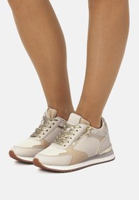 Beige-weiße Sneakers aus strukturiertem Stoff mit goldenen Akzenten und einem seitlichen Reißverschluss. Sie haben flache Schnürsenkel und eine runde Zehenform mit einer Gummisohle.