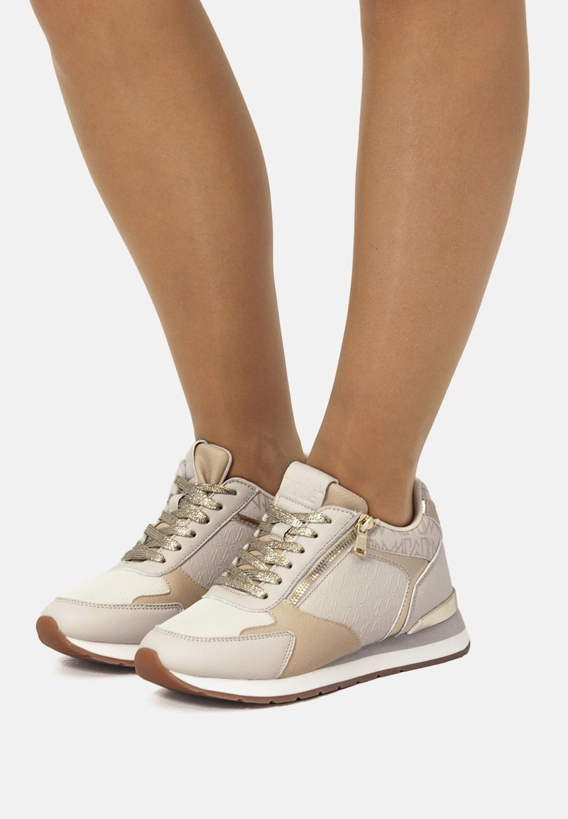 Beige-weiße Sneakers aus strukturiertem Stoff mit goldenen Akzenten und einem seitlichen Reißverschluss. Sie haben flache Schnürsenkel und eine runde Zehenform mit einer Gummisohle.