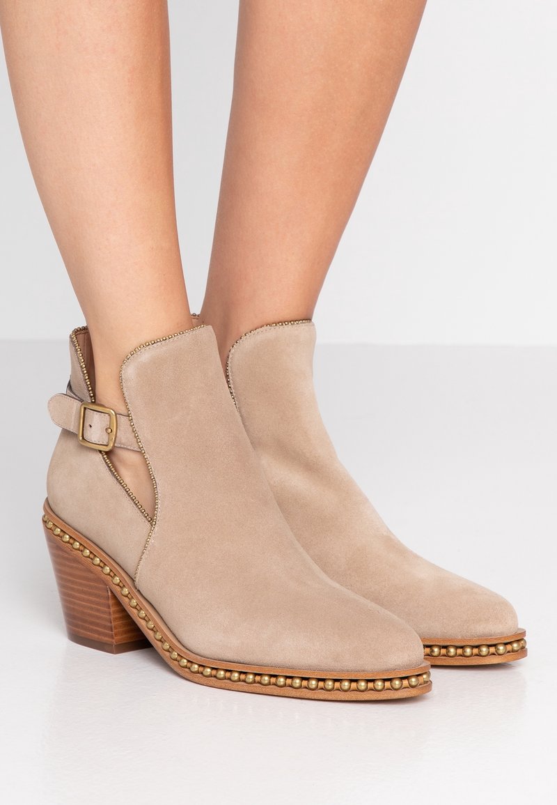 Beżowe zamszowe botki za kostkę z złotymi ćwiekami wzdłuż podeszwy, na blokowym obcasie, z bocznym zapięciem, noszone przez osobę na białym tle.