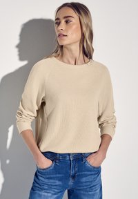 Felpa beige a costine con maniche raglan, vestibilità rilassata. Indossata con jeans di denim blu, mani nelle tasche, ombra sul muro sullo sfondo.