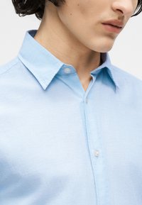 Κοντινό πλάνο ενός ατόμου που φοράει ανοιχτό μπλε πουκάμισο με κουμπιά και αιχμηρό γιακά, εμφανίζοντας τον λαιμό και το κάτω μέρος του προσώπου.