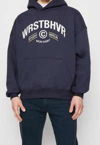 Sudadera con capucha azul marino y un bolsillo frontal, con texto en blanco y amarillo: "WRSTBHVR STUDIO BERLIN MON HABIT." Textura de tela suave.