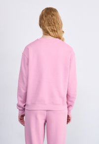 Sweatshirt rose avec une coupe décontractée, des poignets et un ourlet côtelés, et une texture douce. La vue arrière montre un tissu lisse et un design simple.