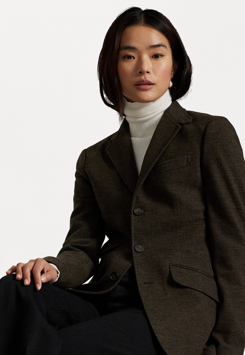 Dark brown blazer made from textured fabric, featuring a notched collar, two buttons, and a single chest pocket, layered over a white turtleneck.
