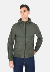 Giacca leggera con cappuccio color verde oliva, chiusura frontale con zip, tasche laterali, polsini e orlo a costine, texture liscia, design casual.