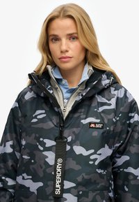 Kurtka w kamuflażu w ciemnoniebieskich i szarych odcieniach, wykonana z materiału wodoodpornego, z zapięciem na zamek i metką z logo. Podszewka ze szarego materiału.