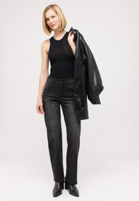 Schwarzes ärmelloses Oberteil, dunkle Jeans, schwarze Lederjacke über dem Arm, und schwarze spitze Absatzschuhe. Glatte Textur, figurbetonter Schnitt.