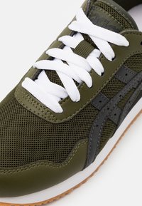 Olivgrön sneaker med nät- och lädermaterial, vita snören och en svart logotyp. Gummisula med texturerat greppmönster.