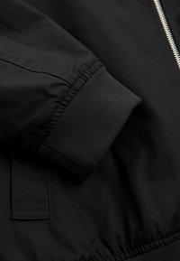 Manche de veste noire avec poignet élastique, poche latérale visible et partie d'une fermeture éclair argentée à l'avant.
