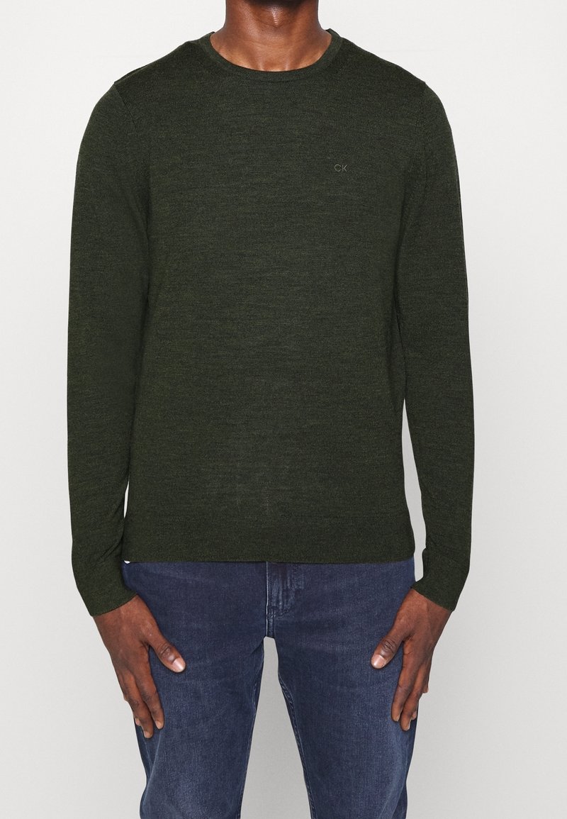 Homme portant un pull en maille à manches longues vert foncé avec un petit logo "CK" sur la poitrine, associé à un jean bleu, debout devant un fond uni.