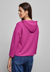 Sudadera fucsia con un patrón texturizado, corte holgado, mangas largas y capucha, combinada con unos vaqueros de denim azul.