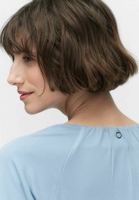 Lyseblå bluse med en glat tekstur, der har en rund knapdetalje på bagsiden og bløde folder langs halsudskæringen.