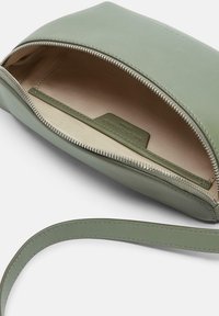 Zielona skórzana torba z zamkiem błyskawicznym, kremowym wnętrzem i paskiem na ramię. Charakteryzuje się gładką fakturą i subtelnym logo wewnątrz.