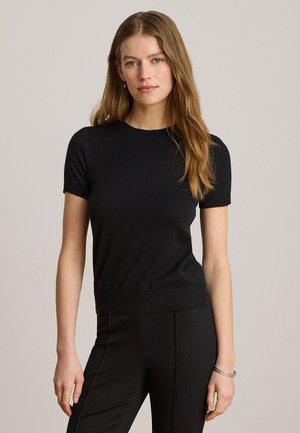 Giovane donna con lunghi capelli castano chiaro che indossa una maglia nera aderente a maniche corte e pantaloni neri, in piedi davanti a uno sfondo neutro.