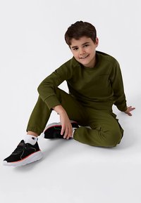Bambino seduto per terra che sorride, indossando una felpa verde e pantaloni con sneakers bianco e nere con rifiniture rosse.