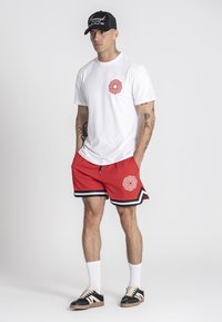 Biela tričko s červeným vzorom, červené šortky s čiernym a bielym lemovaním, biele ponožky a čierna čiapka; s teniskami s béžovými a čiernymi pruhmi.