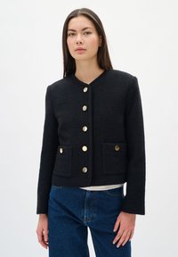 Žena nosi crni teksturirani sako sa zlatnim dugmadi i dva prednja džepa, u kombinaciji sa plavimfarmerkama na bijeloj pozadini.