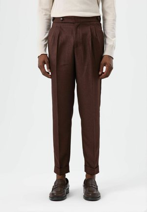 Pantalon en laine marron à coupe ajustée, doté de boutons doubles, de plis sur le devant et d'ourlets retroussés. Porté avec des mocassins foncés.