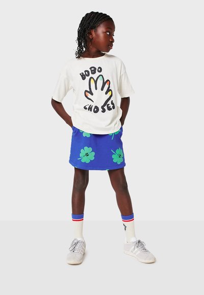 Vit t-shirt med färggrafik av en hand och text, kombinerad med blå kjol med gröna klövermönster. Grå sneakers och vita strumpor.