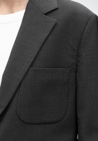 Gros plan sur le revers sombre et la poche poitrine d’un blazer gris foncé porté sur un t-shirt blanc à col rond.