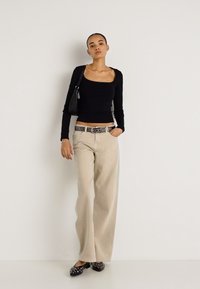 Juodas ilgomis rankovėmis viršus, vienspalvės smėlio spalvos plačios kelnės su raštuota juosta, juoda rankinė ir juodi batai su smeigtukais neutralaus fono fone.