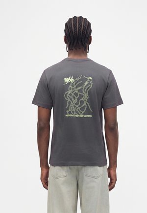 Mand med fletninger iført en mørkegrå t-shirt med gul grafik og teksten "Never Stop Exploring" på ryggen.
