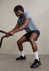 Gray short-sleeve athletic shirt, black shorts, black and gold sneakers, and white socks; using battle ropes in a neutral backdrop.