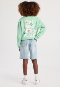 Felpa oversize verde menta con testo bianco e adesivi con logo sulla schiena, abbinata a pantaloni denim azzurri e sneakers bianche.