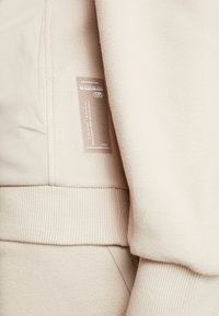 Beige Fleecejacke mit gerippten Bündchen und einem Logosticker im Innenbereich. Glatte Textur mit einem weichen, warmen Erscheinungsbild.