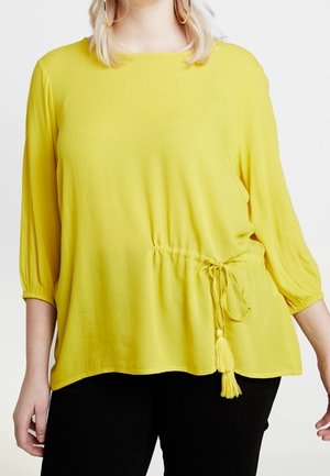 Nainen, joka pukeutuu kirkkaan keltaiseen puseroon, jossa on 3/4-hihat ja hapsunaru vyötäröllä, yhdistettynä mustiin housuihin.