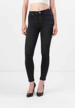 Vrouw draagt zwarte high waist skinny jeans en zwarte lakleren hoge hakken, staand tegen een effen witte achtergrond.