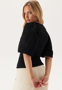 Top nero con texture, maniche corte e voluminose, vita aderente e scollo tondo. Il materiale appare stropicciato, aggiungendo profondità e dimensione.