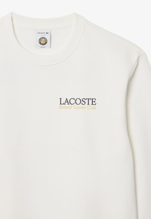 Camiseta blanca de manga larga con cuello redondo, texto "LACOSTE Roland Garros Club" en el pecho y una etiqueta de marca dentro del cuello.