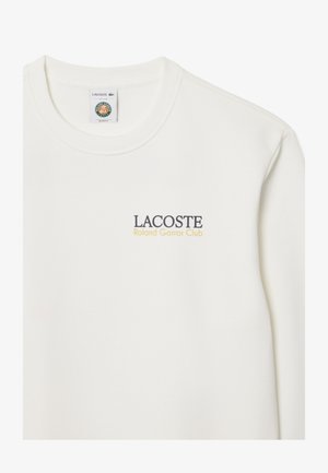 Camiseta blanca de manga larga con cuello redondo, texto "LACOSTE Roland Garros Club" en el pecho y una etiqueta de marca dentro del cuello.