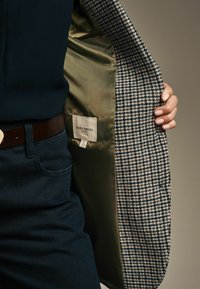 Blazer mit einem Hahnentrittmuster, grauer und marineblauer Außenansicht, olivgrüne Satinfütterung und einem Etikett im Inneren. Verfügt über eine taillierte Passform und eine strukturierte Form.
