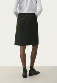 Gonna in corda verde scuro con vita elastica, lunghezza al ginocchio e texture liscia. Indossata con scarpe nere e calze.