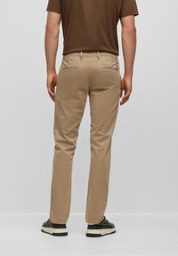 Beige chino kalhoty s úzkým střihem, se dvěma zadními kapsami a hladkým textilním povrchem, v kombinaci s černými teniskami na světlém pozadí.