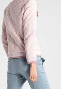 Person in einer hellrosa gesteppte Jacke und hellblauen Hosen, von hinten und zur Seite sichtbar vor einem schlichten Hintergrund.