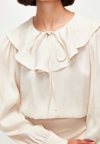 Blusa crema con un ampio colletto a strati, chiusura con nastro al collo e maniche lunghe con polsini arricciati, realizzata in un tessuto leggero e testurizzato.