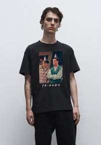 Černé tričko s grafikou ztvářující dvě postavy v party kloboucích z televizního pořadu, obklopené textem "F.R.I.E.N.D.S."